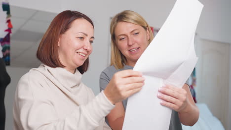designers smiling and laughing together while examining paper design pattern during discussion, joyful atmosphere in bright creative workspace, highlighting teamwork and professional environment