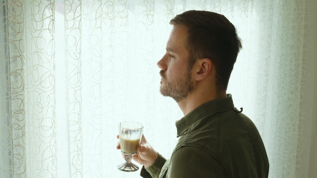 A man looks out of the window and drinks a glass of milk. the sun shines through the curtains