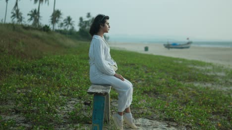 woman in white sitting on a bench by the beach at daylight, looking thoughtful, with a boat and sea in the background, serene