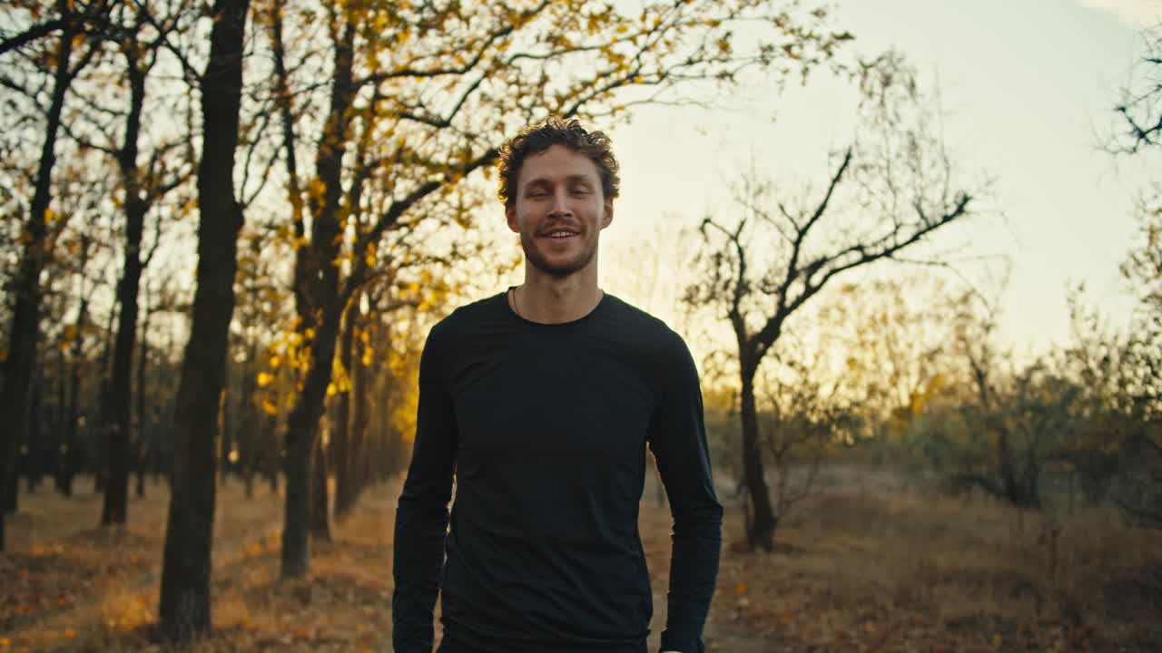 Portrait of a man with curly hair in a black sports uniform who looks confidently at the camera and smiles while holding his arms crossed on his chest In the autumn forest in the morning