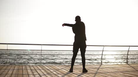 Young-sporty-man-stretching-out-on-the-dock-near-the-seaside.-Beautiful-sunny-morning-on-the-seacoast.-Outdoors-footage.-Backside-video.