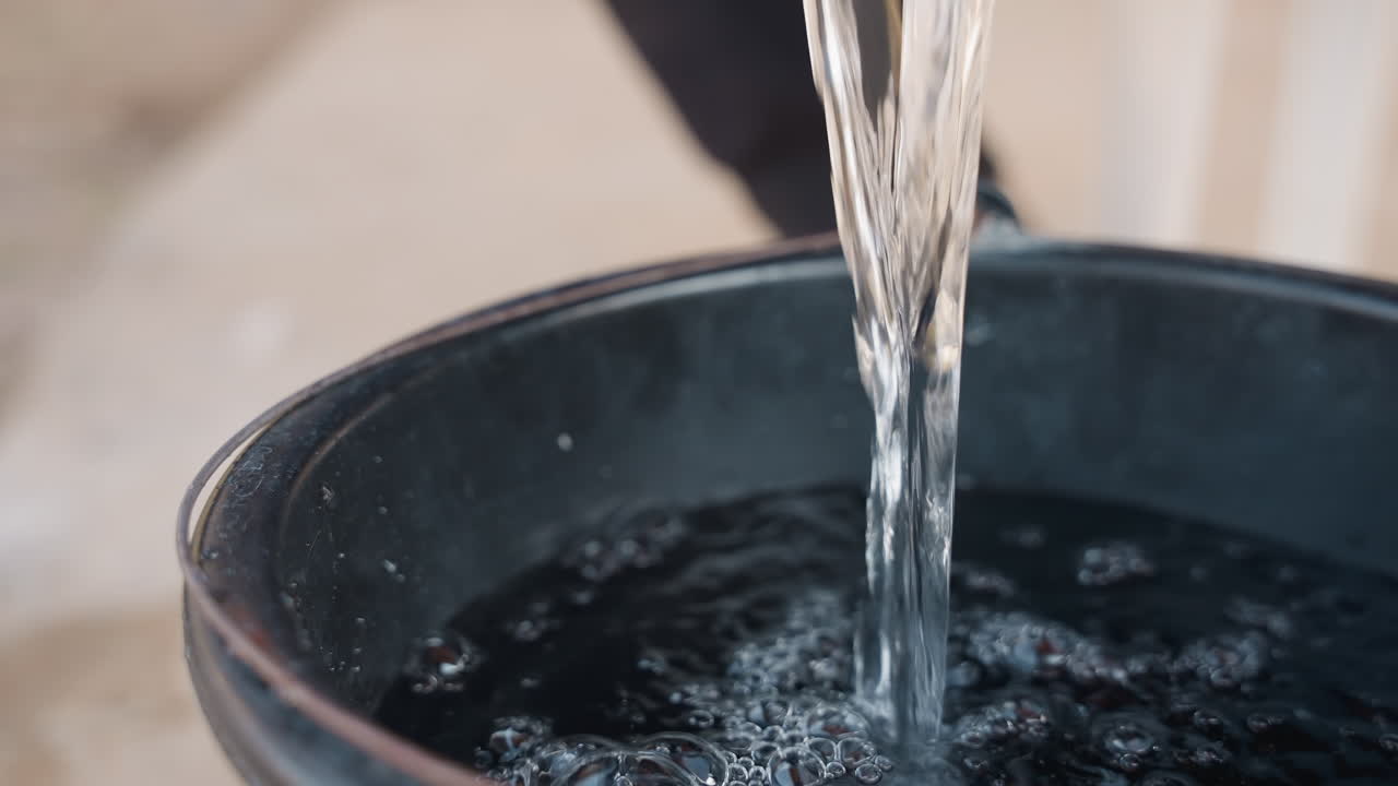 Falegname che versa acqua in un secchio presso la stazione di lavaggio dell'officina, braccio e manica visibili, un getto vigoroso che colpisce il secchio scuro producendo bolle e schizzi, sfondo di cemento grezzo e pulizia degli attrezzi