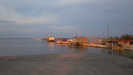 Vista-Aérea-De-Cabañas-De-Pesca-A-Orillas-Del-Estuario-Al-Atardecer,-Máquina-De-Pesca-Italiana,-Llamada-&quot