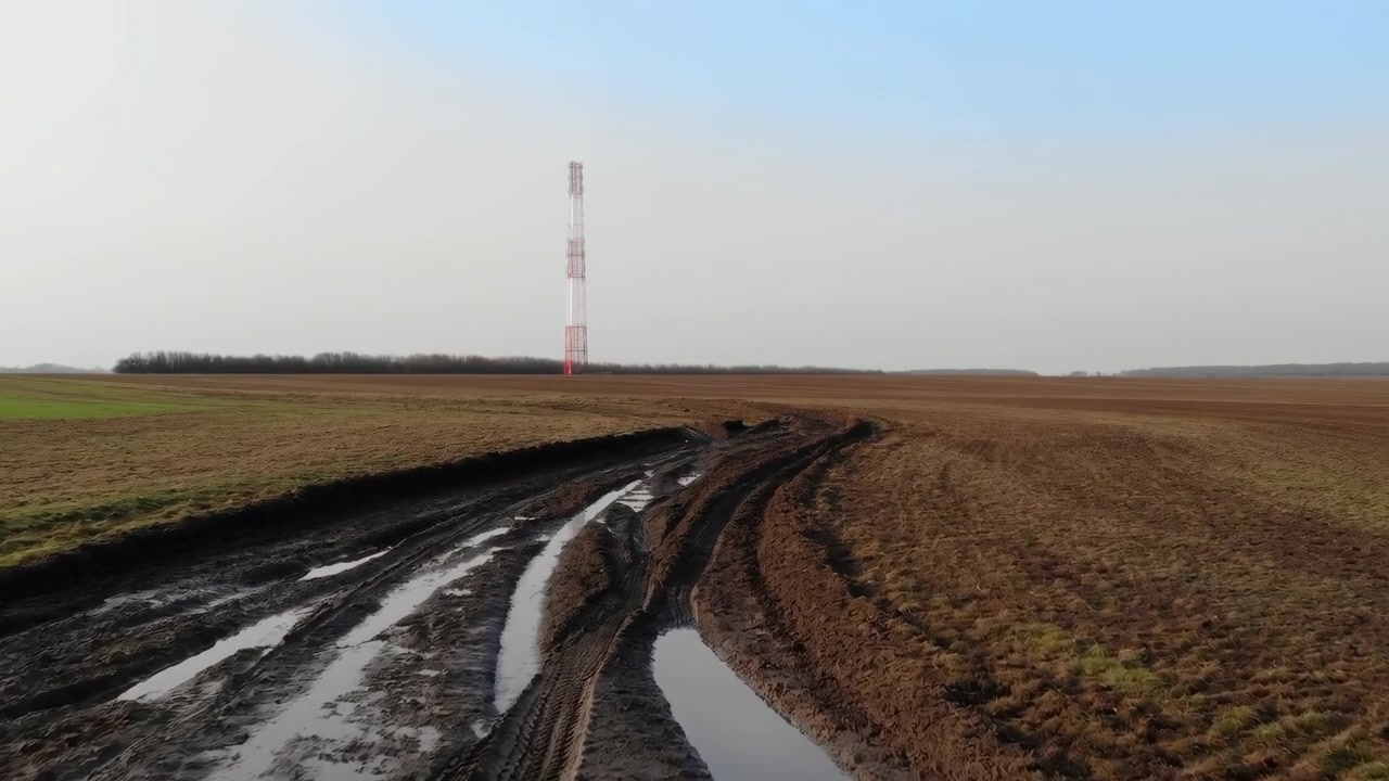 Solchi profondi e fangosi di trattore, pieni di acqua stagnante riflettente, si curvano attraverso il primo piano e guidano lo sguardo attraverso un ampio campo incolto verso un'isolata torre di comunicazione a traliccio bianca e rossa sull'orizzonte basso.