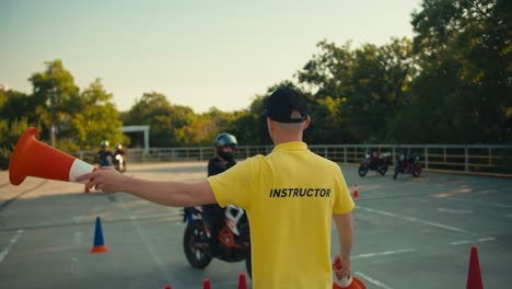 a driving instructor in a yellow t-shirt indicates to a motorcyclist which way he should turn with the help of orange cones