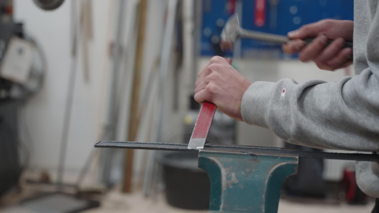 Close-up handheld of young apprentice man hands working with hammer and a red chisel on steel in a blacksmith workshop