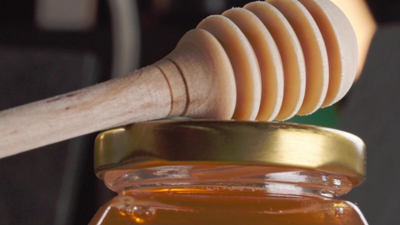 Mestolo di legno per miele appoggiato sopra un barattolo di vetro riempito di miele dorato. Il primo piano evidenzia il manico di legno scanalato, la superficie liscia del miele e il contenitore di vetro trasparente.
