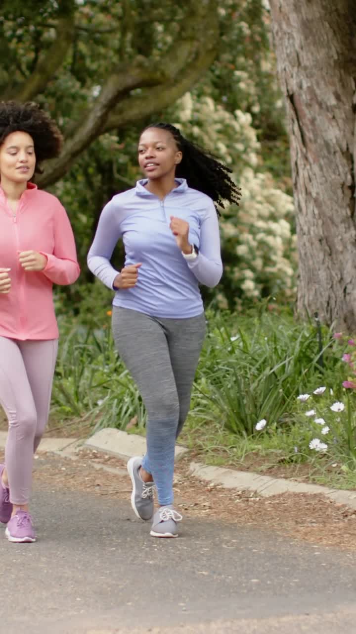 Video verticale: Due donne che fanno jogging su un sentiero del parco oltre un albero, indossando maglie rosa e blu, con un orologio.