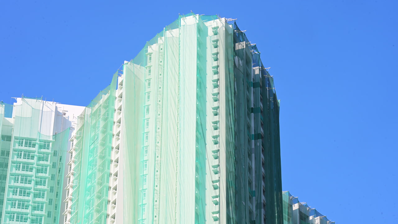 Close-up of a high-rise residential facade draped in green safety netting for renovation in Hong Kong. Establishing shot of urban building maintenance and real estate refurbishment in China.