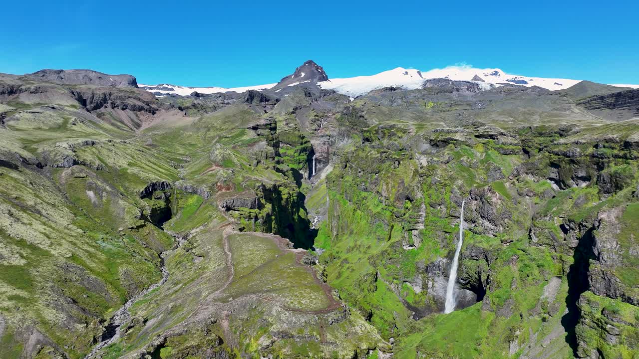 Aerial rising shot of green steep and rocky Mulagljufur canyon with waterfall and snowy peak. Sunny summer day with blue sky