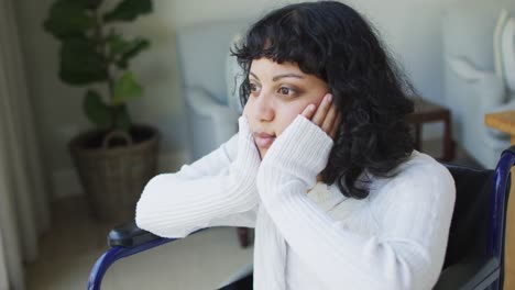 Smiling-biracial-disabled-woman-in-wheelchair-looking-out-of-window-in-living-room