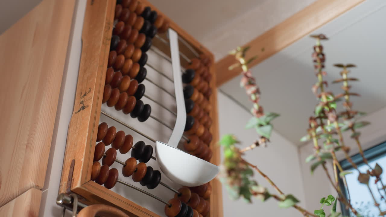 Close up of flower stems in soft focus foreground with white kitchen ladle hanging on wooden vintage abacus in bright corner near window, combining traditional decor with natural greenery