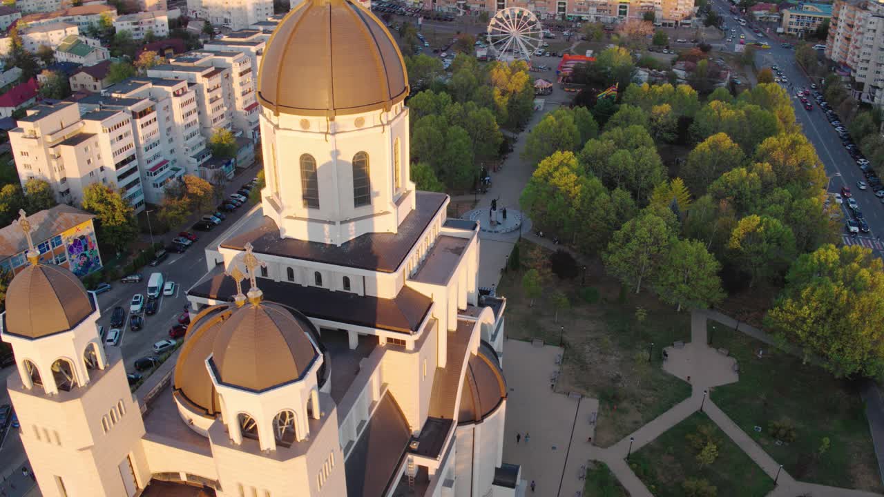 A cinematic aerial shot slowly orbits the main dome of a majestic Orthodox cathedral in Bacau, Romania, during a beautiful golden hour sunset, revealing the cityscape