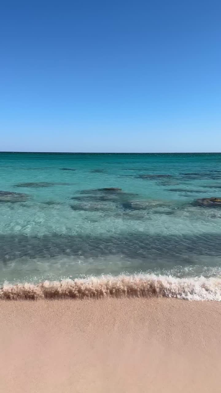 Veduta panoramica verticale di onde leggere che lambiscono una spiaggia sabbiosa con acqua cristallina color turchese e orizzonte calmo lungo la costa del Mar Egeo in Grecia.