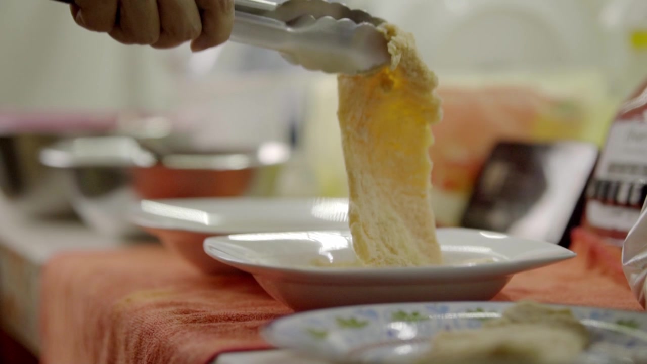 Close-up of a hand lifting a piece of cooked chicken with metal tongs over a white plate. The food is being prepared and served at a dining table indoors