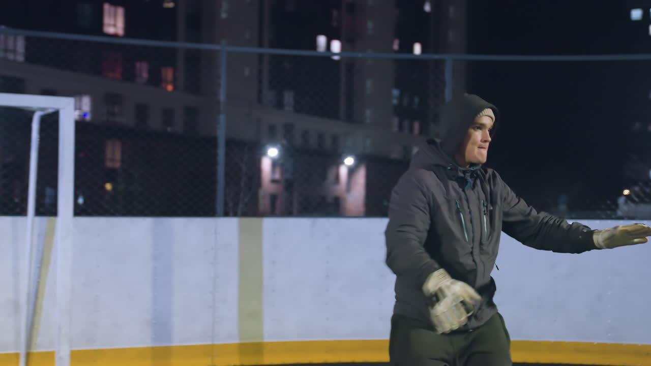 Man in hoodie kicks soccer ball near goalpost during evening training session on urban outdoor field, shadow reflections and illuminated background buildings