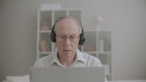Ein-älterer-Mann-Mit-Kopfhörern-Spricht-Und-Schaut-Sich-Die-Anzeige-Seines-Notebooks-An,-Während-Er-Online-Von-Zu-Hause-Aus-Vorlesungen-Hält