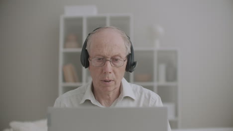 un anciano con auriculares está hablando y mirando la pantalla de un cuaderno que da conferencias en línea desde casa
