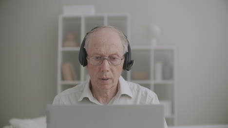 elderly man with headphones is speaking and looking at display of notebook lecturing online from home