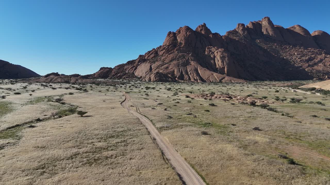 Scenic drone view of Spitzkoppe mountain range in Namibia, surrounded by golden desert sands and expansive rock formations under a blue sky with off road path
