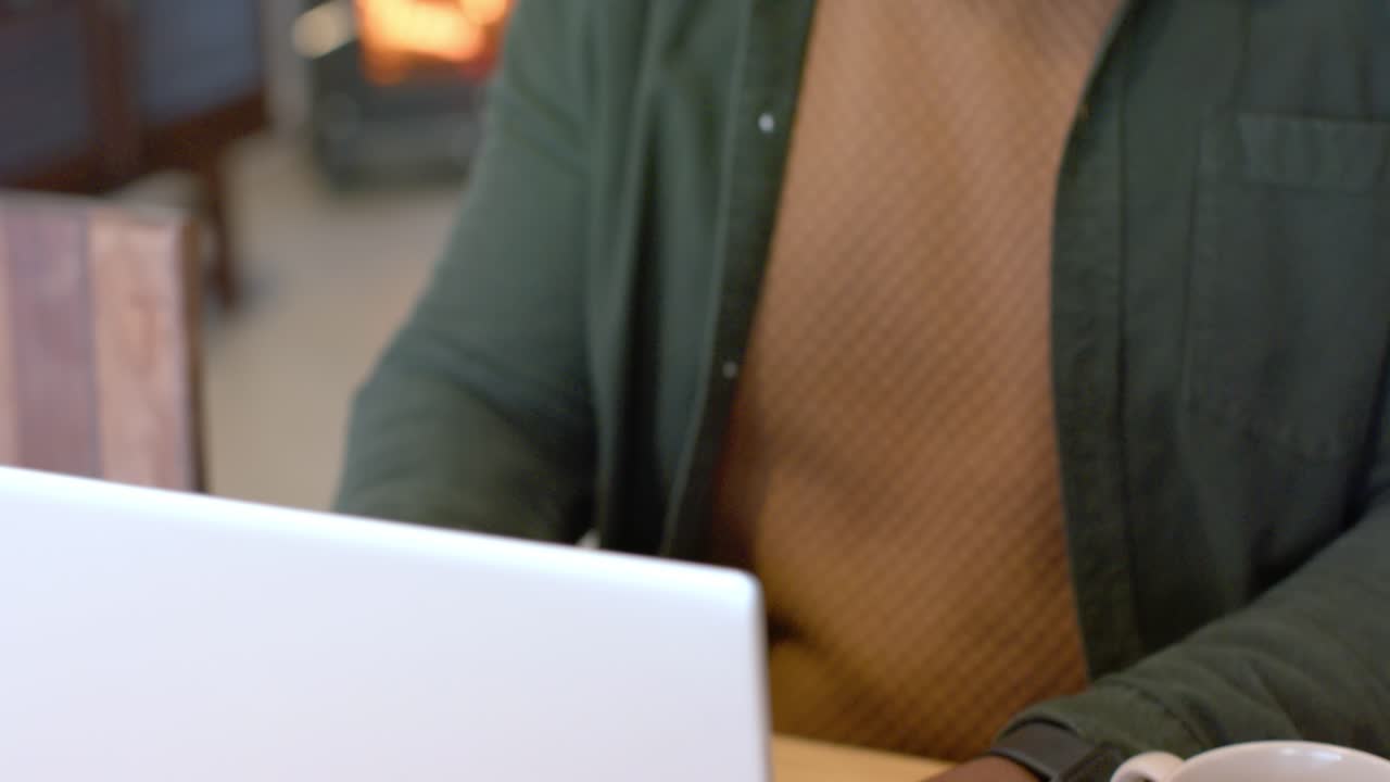 Camera pulling back from dark hands typing on silver laptop for work, revealing face near fireplace