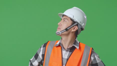 close up of asian male engineer wearing safety helmet looking around while standing with arms akimbo in the green screen background studio