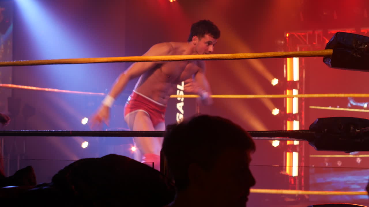 Two wrestlers engaged in a fast-paced move inside a brightly lit wrestling ring, with an intense atmosphere created by dramatic stage lighting. Surrounded by a captivated audience