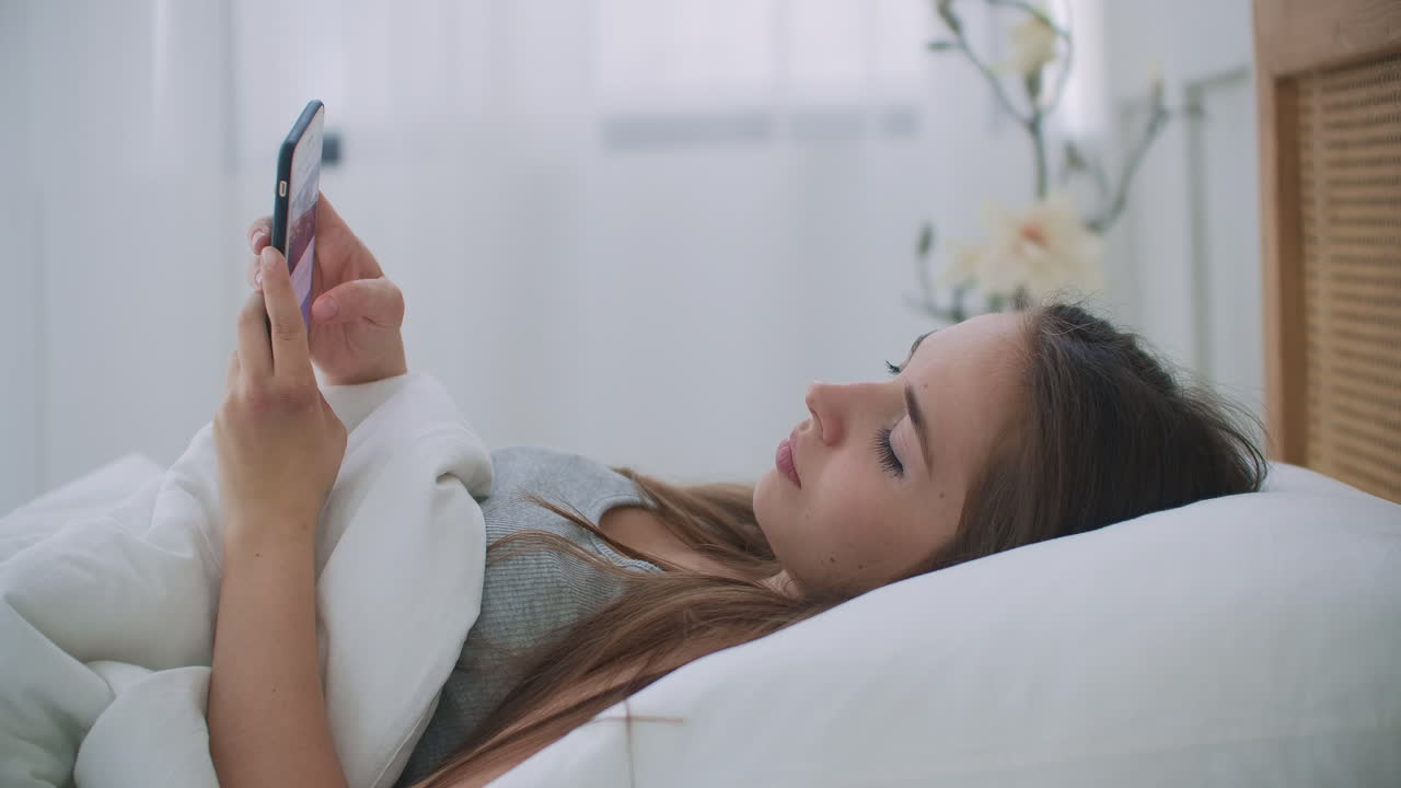 집에서 침대에 누워있을 때 휴대폰을 들고 있는 여성.