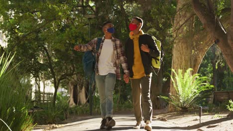 two happy mixed race gay male couple walking and talking in park with backpacks