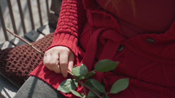 Primo piano di una donna con un maglione rosso che tiene un ramo verde frondoso su una panchina esterna con ringhiera in metallo e cuscino in maglia marrone. La luce del sole proietta ombre, enfatizzando i dettagli della texture come la trama del tessuto.