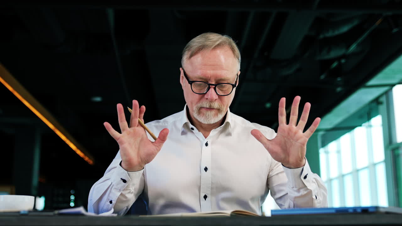 Calm pensive man wearing white shirt and glasses sits at desk. Mam puts away the paper notebook and opens laptop. Low angle view.