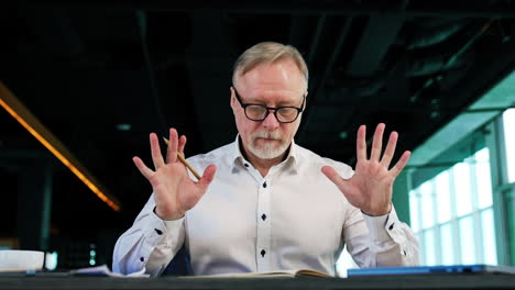 calm pensive man wearing white shirt and glasses sits at desk. mam puts away the paper notebook and opens laptop. low angle view.
