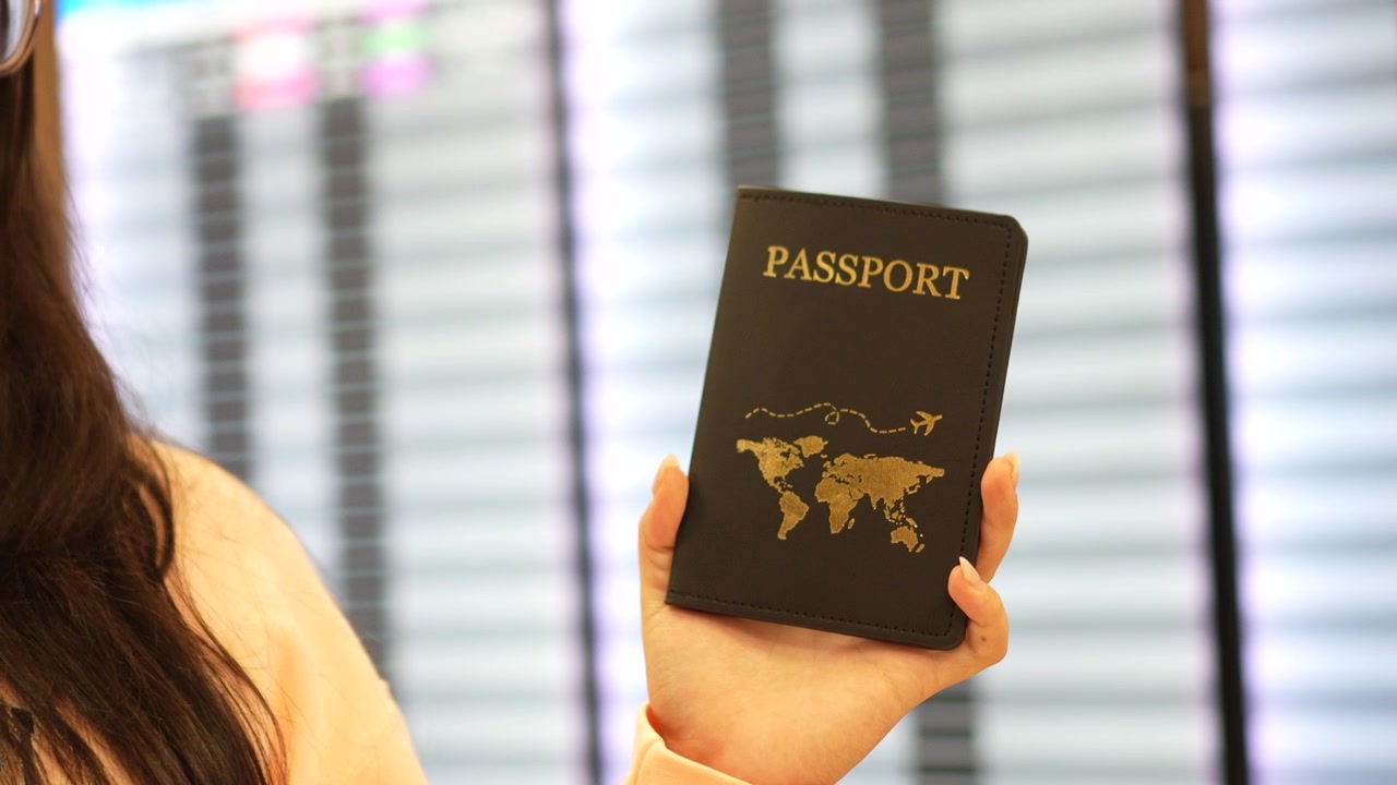 Un primo piano mostra un viaggiatore che tiene un passaporto all'interno di un terminal aeroportuale, con un tabellone delle partenze e degli arrivi sfocato sullo sfondo
