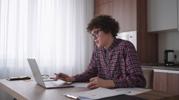 En attraktiv, krølhåret mandlig studerende med briller studerer hjemme. Han bruger sin bærbare computer til at skrive og tage noter i en notesbog. Den studerende ser et online fjernundervisningsseminar på sin bærbare computer.
