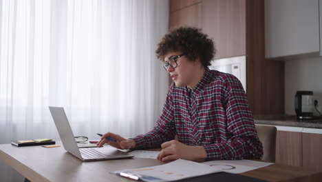 curly haired male student attractive young boy in glasses is studying at home using laptop typing writing in notebook. college student using laptop computer watching distance online learning seminar