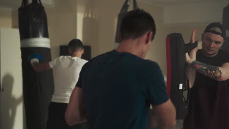 robust men boxers execute proper punching technique using specialized gear and punching bag. group workout. guidance from coach to participants