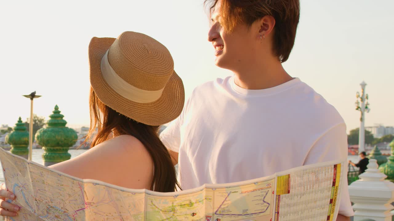 Asian couple consults map outdoors, warm sunset lighting, riverside travel, candid joyful interaction