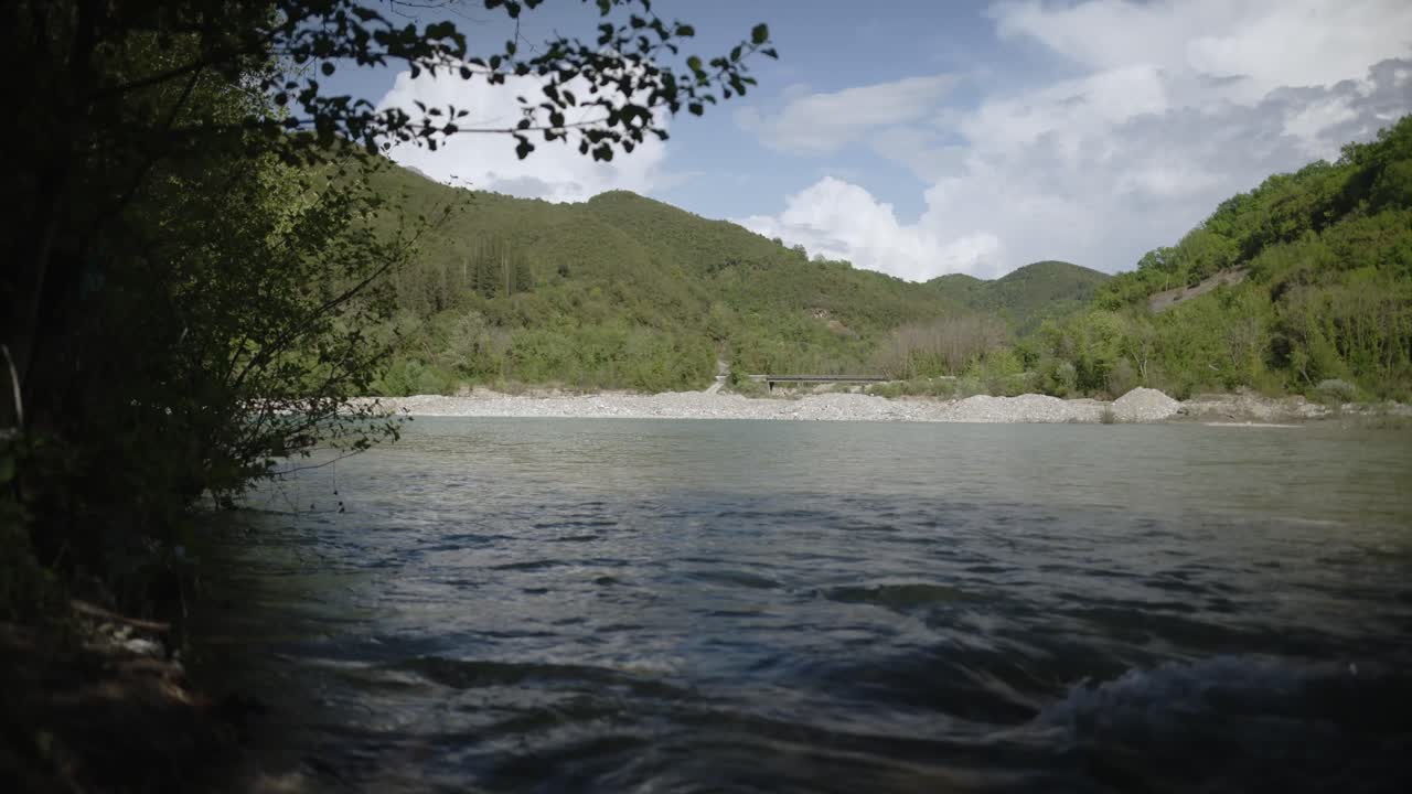 Serene river flows through a forested valley with tree branches creating a natural frame. Hills rise in the distance under a sky of sunlit clouds. Shot in Tzoumerka.