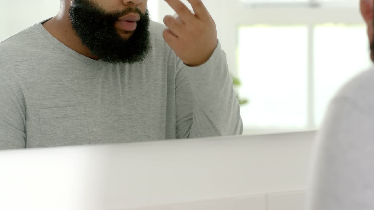Adult African American man blending cream from dots at bathroom vanity mirror, checking skin