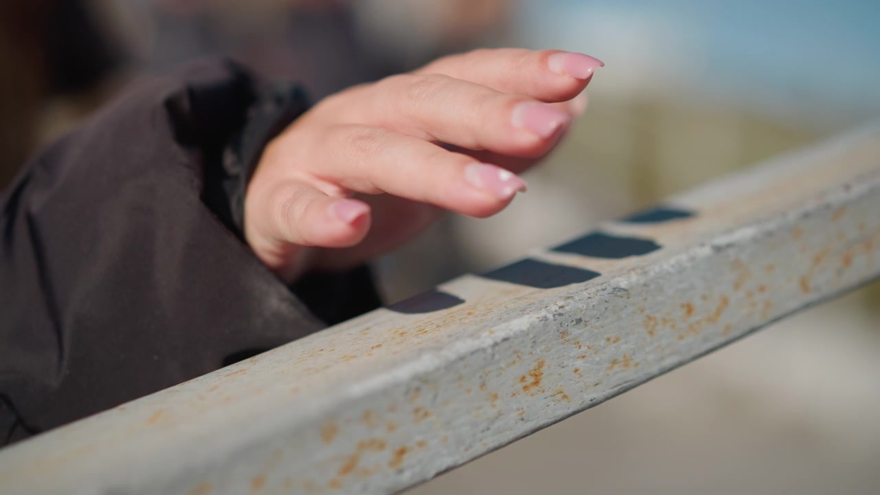 Woman hand with manicured nails rests on weathered metal railing showing subtle movement and texture detail. background is softly blurred suggesting outdoor setting like bridge or deck