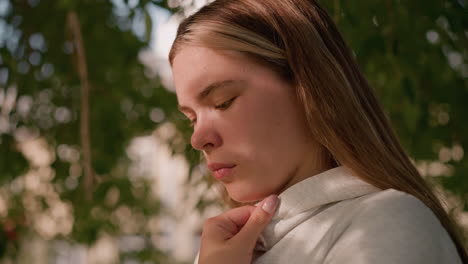 close-up of young woman in casual attire, intensely focused while holding her hood collar, face softly illuminated by sunlight filtering through tree leaves