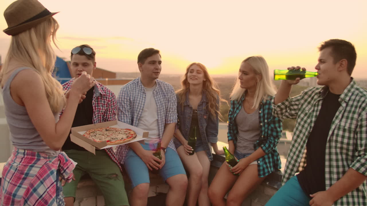 The company of six young people communicate with each other on the roof with pizza and beer. Girl enjoys the smell of hot pizza.