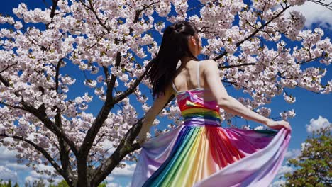 gracefully dancing young woman in flowing rainbow dress twirling under blooming cherry blossoms, capturing vibrant spring elegance against sunny garden backdrop