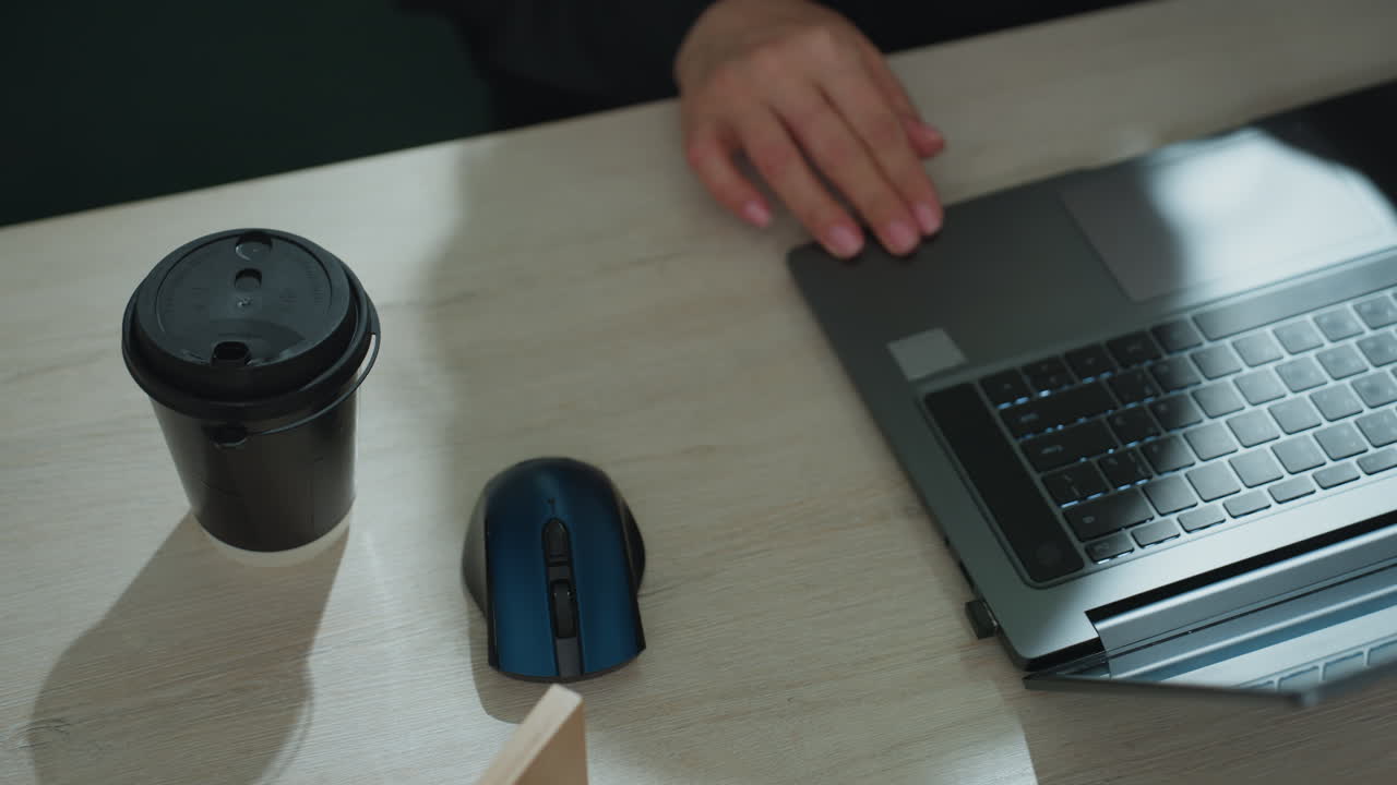 Overhead view of professional woman placing takeaway coffee on desk, hand reaching toward laptop beside wireless mouse and miniature estate model, preparing workstation for focused business activity