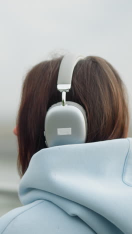 backside view of woman walking and listening to music with headphones, blurred background featuring bridge, water, and iron rail, giving a calm and reflective atmosphere