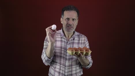 macho adulto con una mirada desconcertada en su rostro examina de cerca un huevo blanco de un cartón de huevos marrones, colocado contra un fondo rojo oscuro