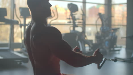 athlete doing exercises in the simulator with weights and pulls the bar sitting on the background of a large window gym. strong purposeful athlete view from the back