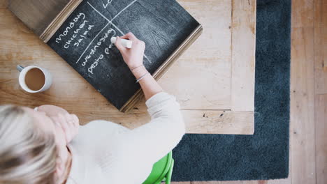 Overhead-Shot-Looking-Down-On-Woman-At-Home-Writing-Meal-Plan-On-Blackboard