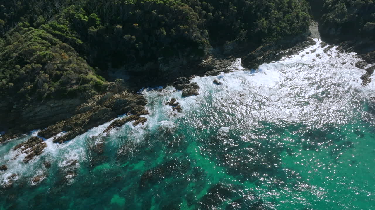 Aerial: Top down birds eye view of the rocky coastline and green blue ocean near Mimosa Rocks, South Coast NSW, Australia