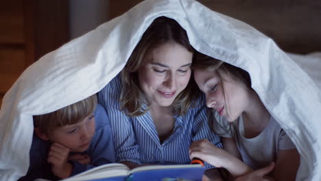 close-up view of mother and her cute little daughter and son using a flashlight and reading a book under the blanket at night